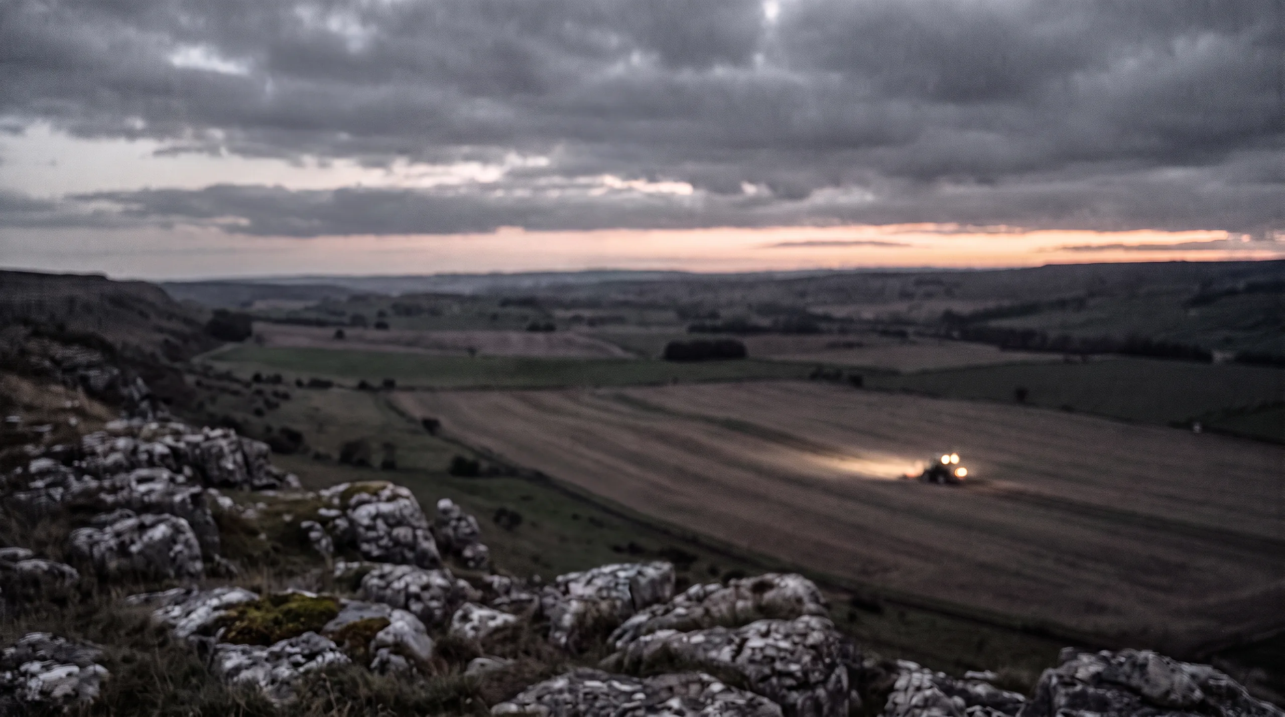 A dusk valley in the west of Ireland with a tractor working straight rows under its lights